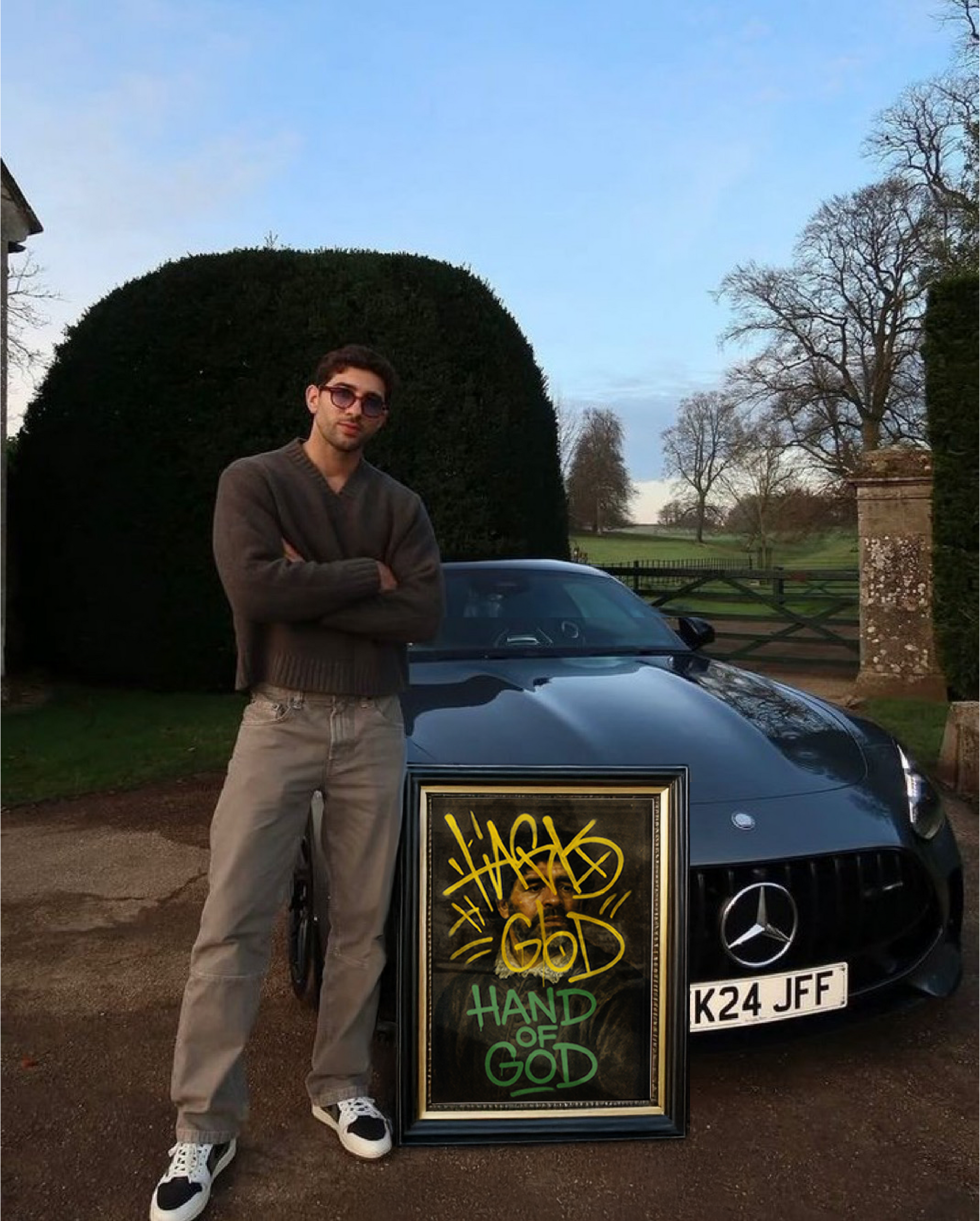 Man standing next to a black Mercedes-Benz car with a framed artwork titled 'Hand of God' in a garden setting.